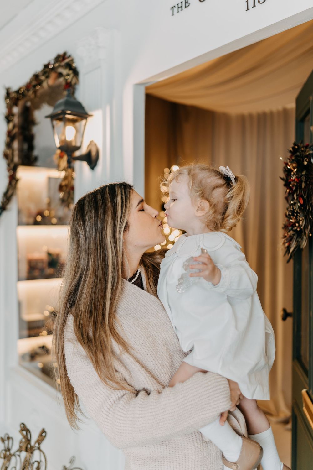 A parent and baby in matching white sweaters share joyful moments in a holiday decorated room.