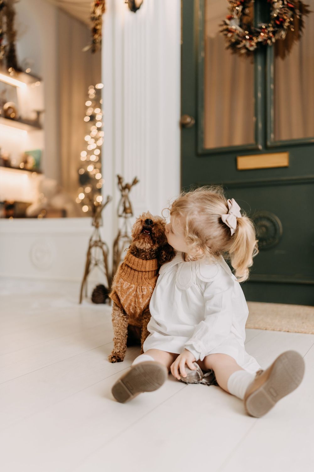 A small child in a white dress sits on the floor with a brown toy dog beside Christmas decorations and twinkling lights.
