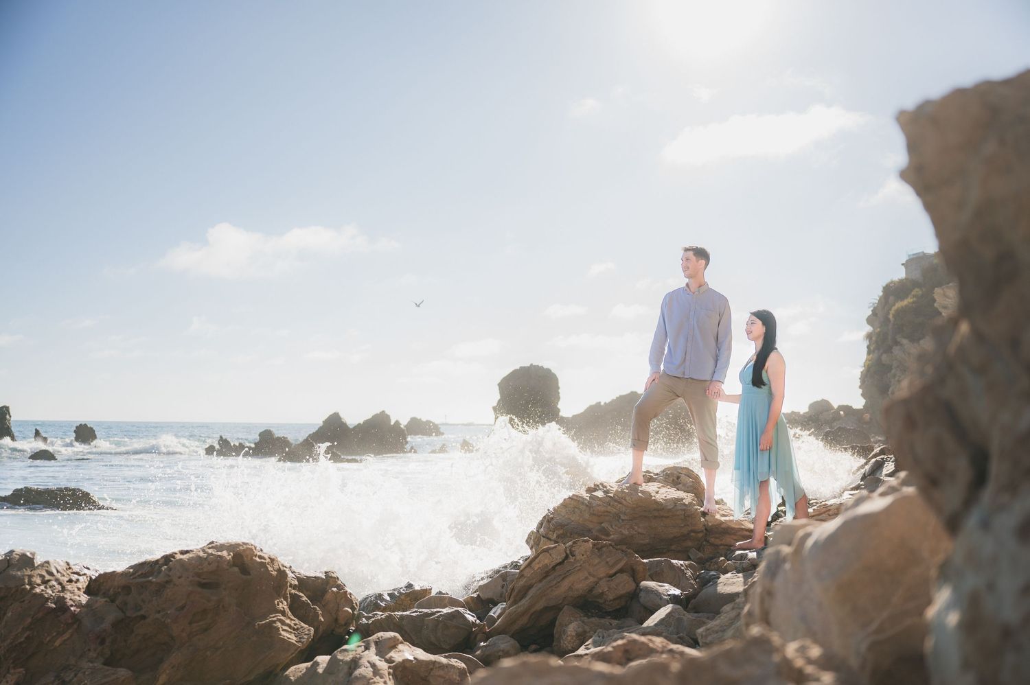 Couple standing on rocky beach during sunset with ocean waves crashing.