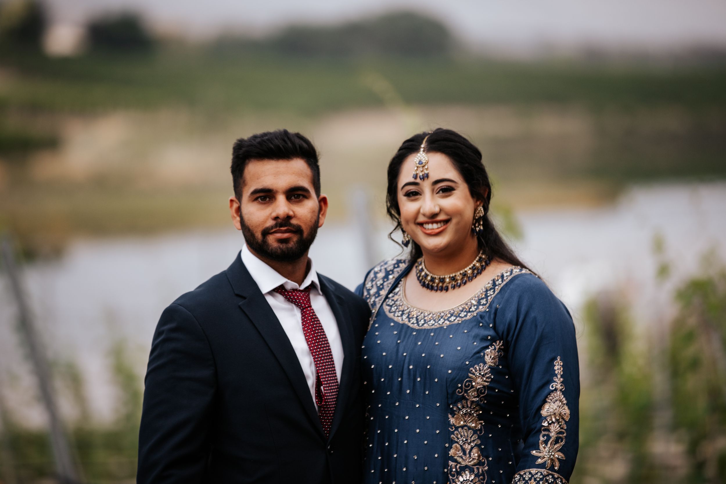 A couple dressed in elegant South Asian wedding attire poses together outdoors near a serene lake.
