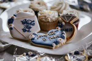 Decorated wedding cookies and cupcakes on a plate, featuring a ring, envelope, and personalized designs.
