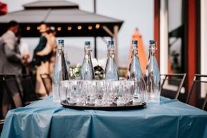 Glass water bottles and drinking glasses arranged on a blue tablecloth at an outdoor wedding reception.
