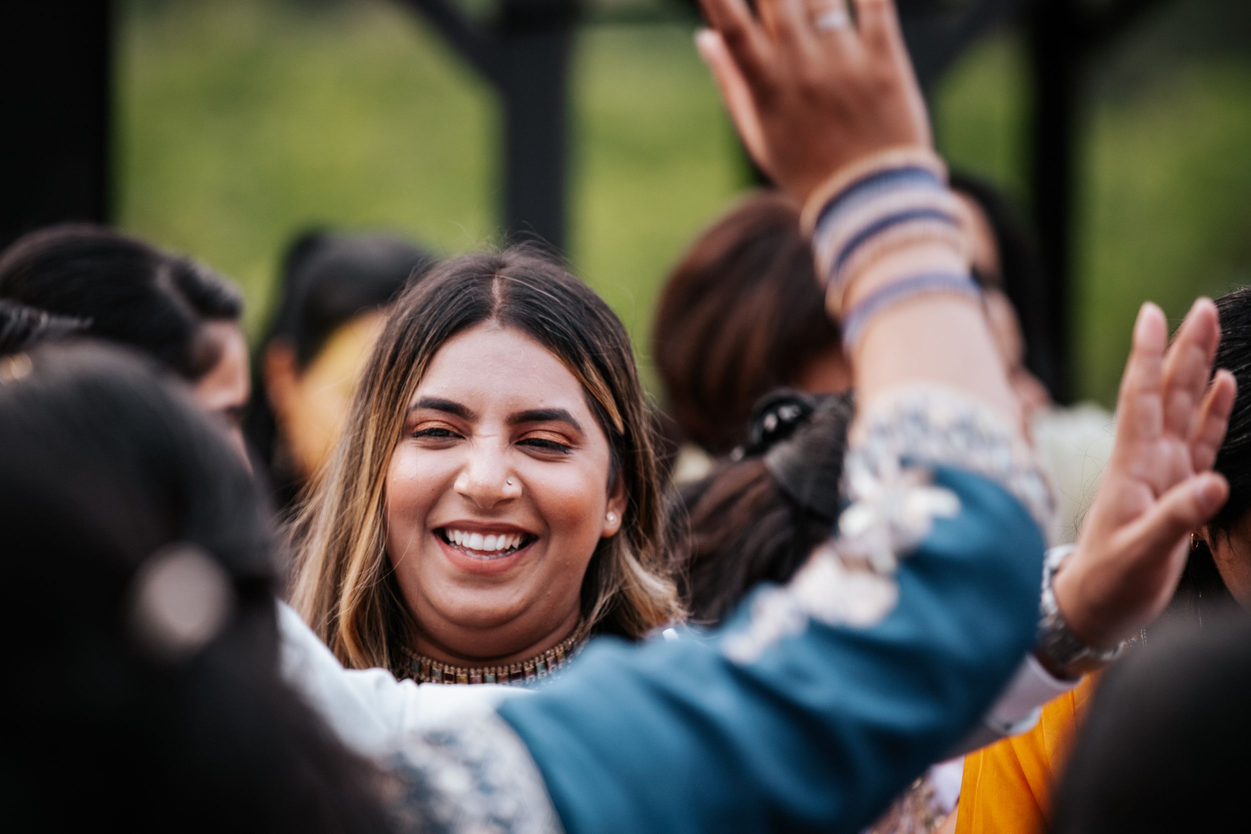 A joyful woman smiles and raises her hand during a lively outdoor wedding celebration.