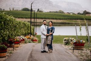 A couple in traditional South Asian wedding outfits poses on a scenic vineyard driveway with a lake view.