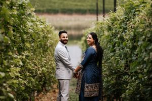 A couple holds hands while walking through lush green vineyard rows on their wedding day.