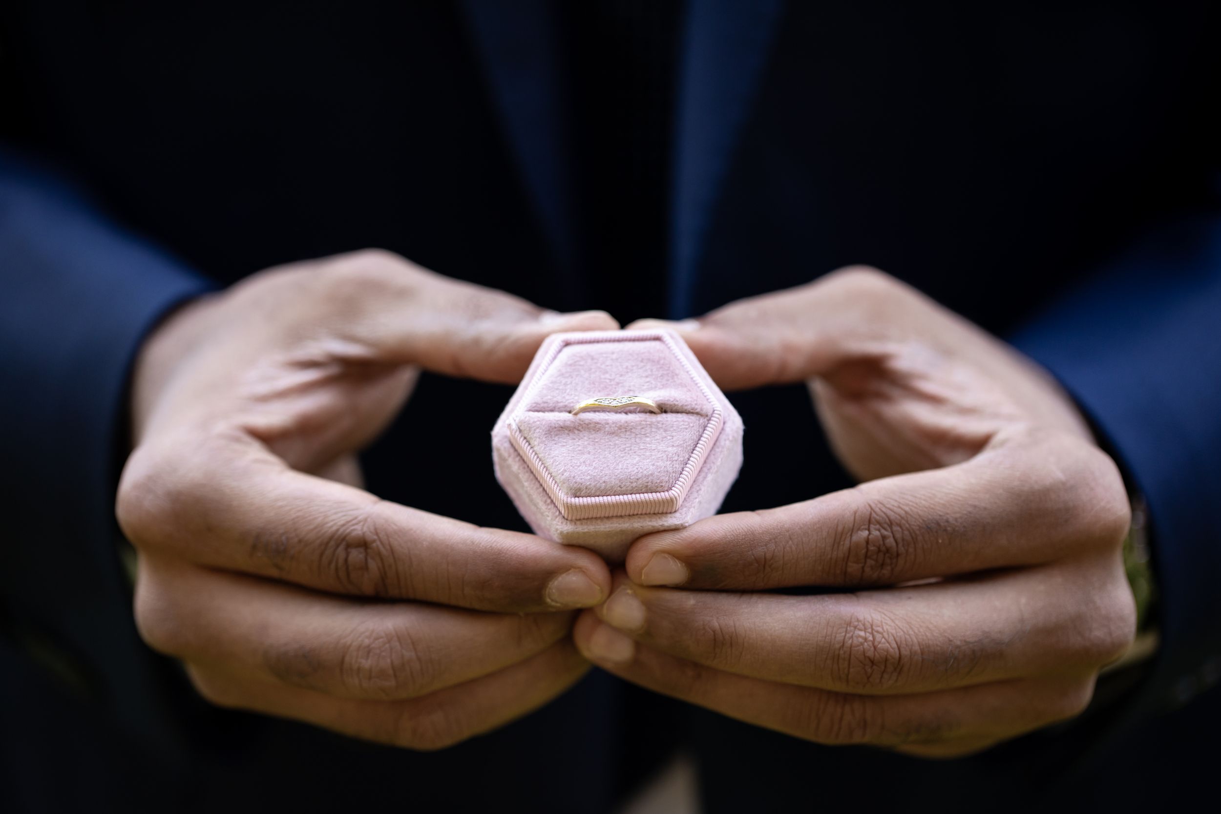 A man in a suit presents a ring in a pink hexagonal velvet box with both hands.