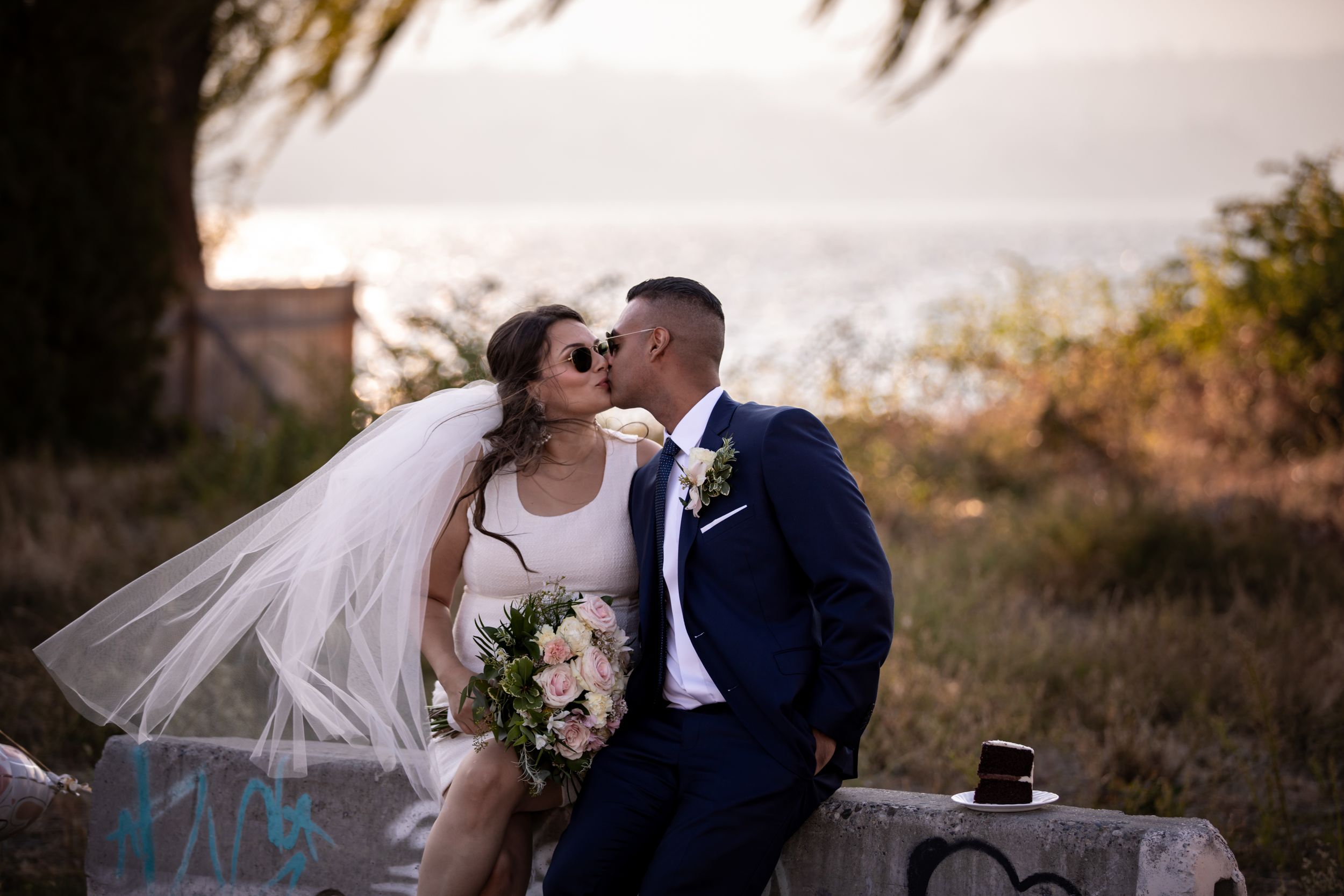 A bride and groom share a romantic kiss outdoors with a scenic ocean view backdrop on their wedding day.