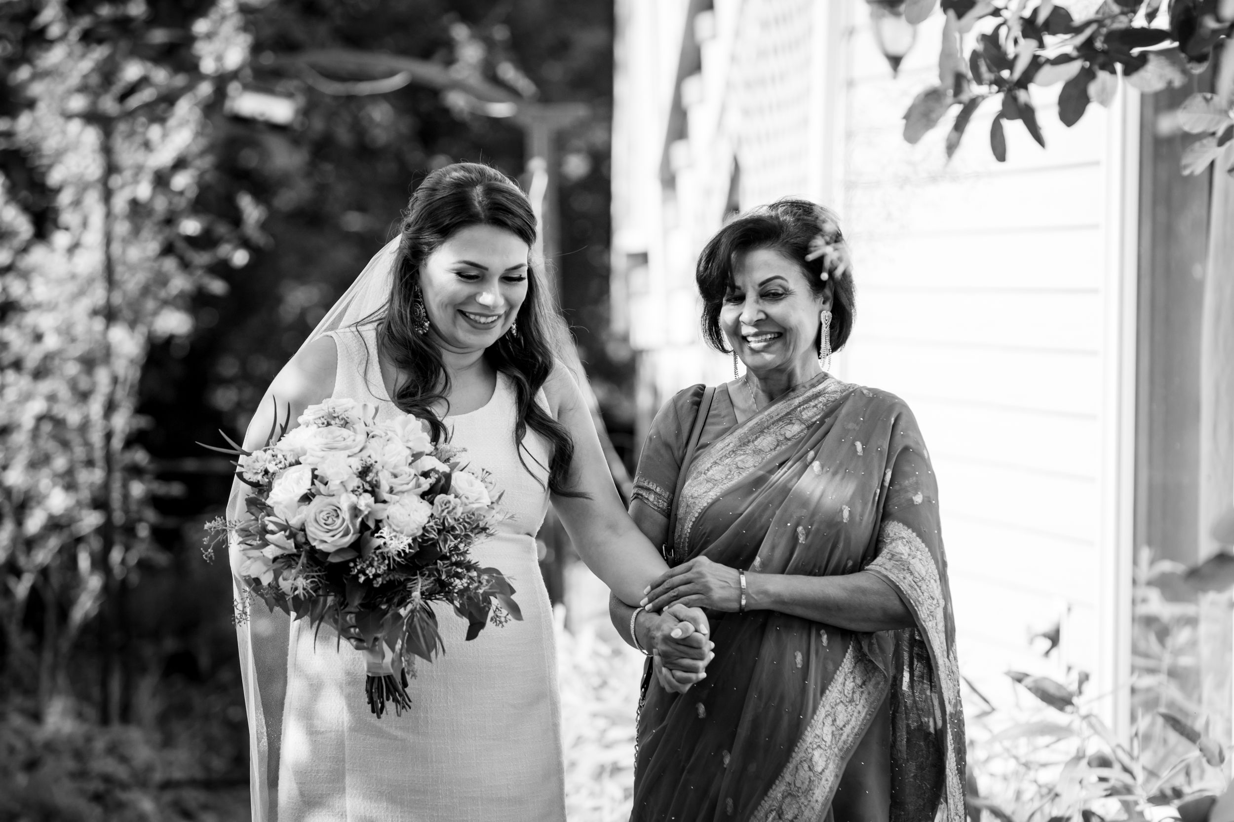 A smiling bride in a white gown and veil walks with an older woman in a sari.
