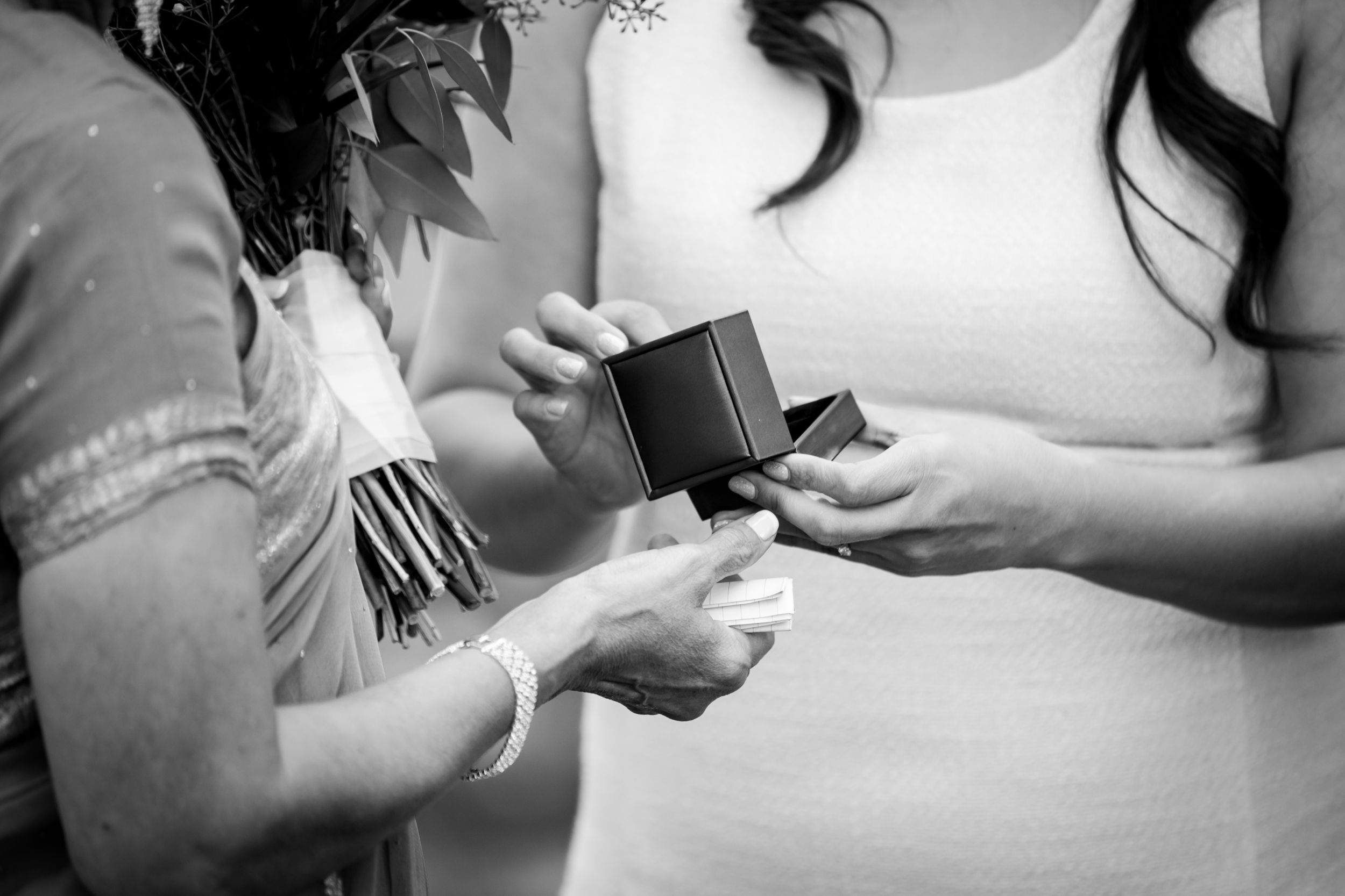 Two women exchange a small black ring box during a wedding ceremony in black and white.