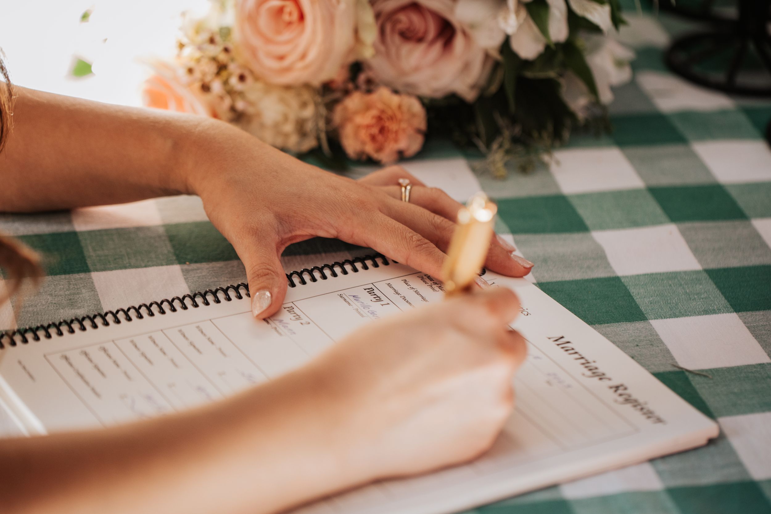 A bride signs a marriage registry document next to a beautiful floral arrangement.