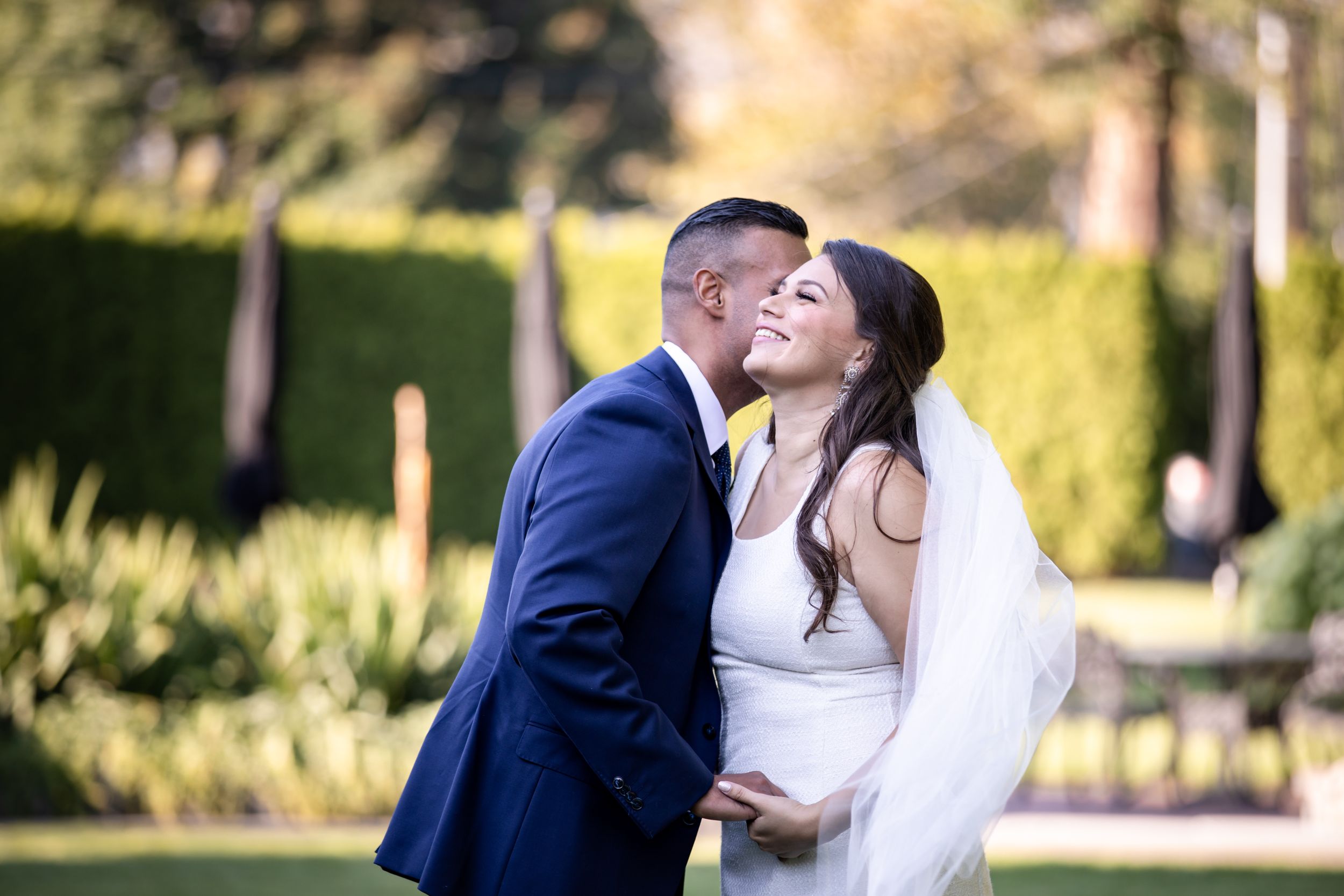 A joyful bride laughs as her groom kisses her cheek in a sunlit garden setting.