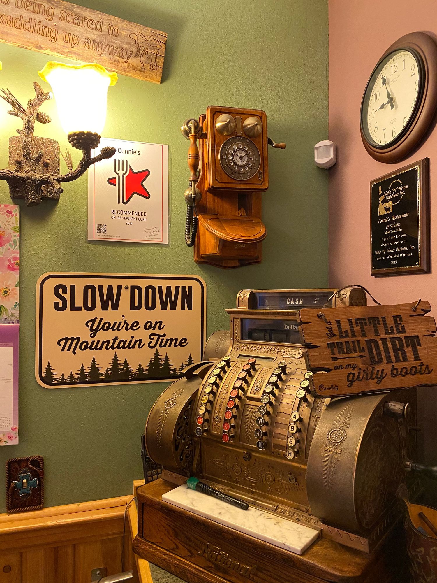 Vintage decor featuring antique cash register, wooden telephone, and rustic mountain-themed sign on green wall.