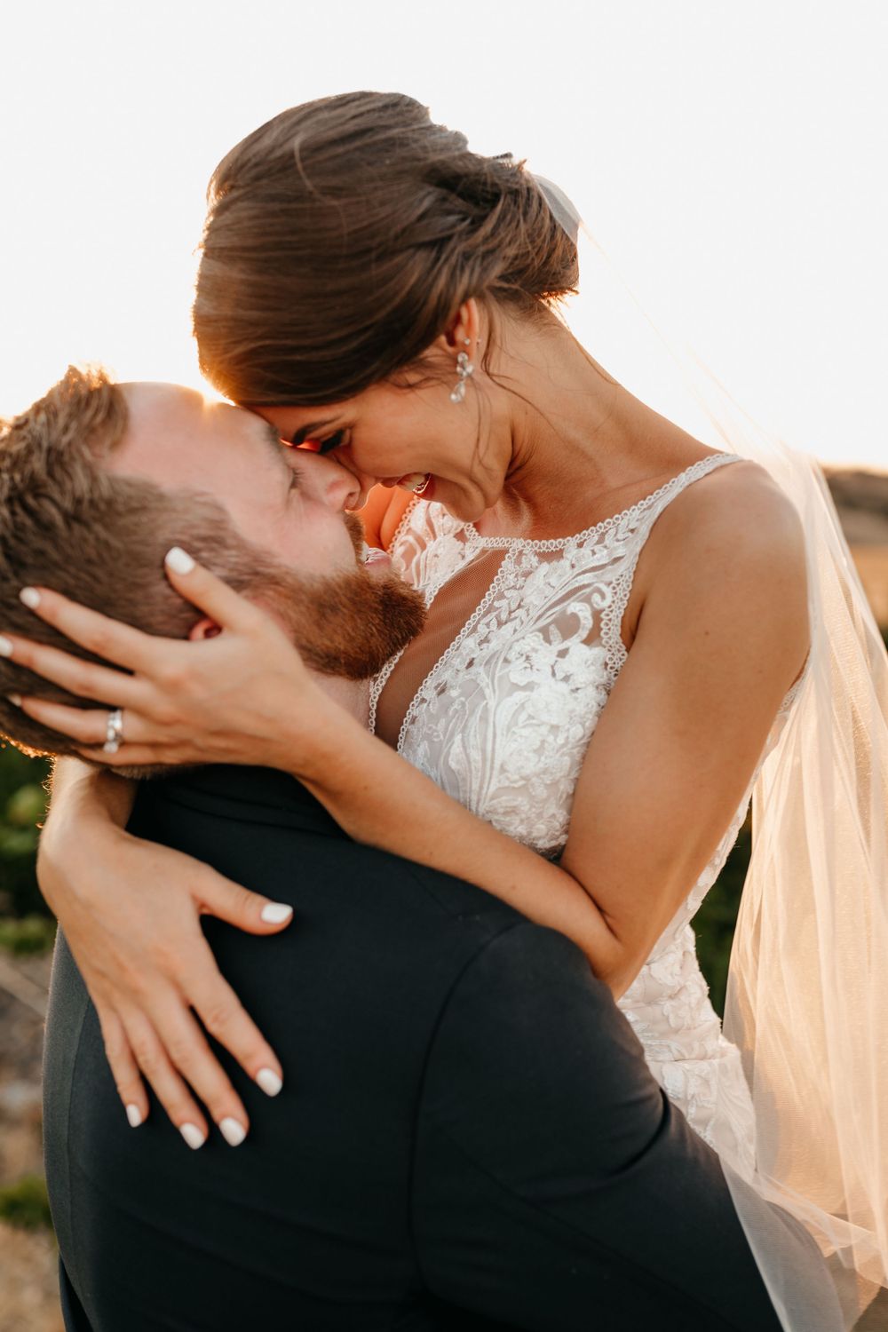 A groom lifts his wife in embrace as she hold his head close to hers. The sun is setting behind them. It is gorgeous.