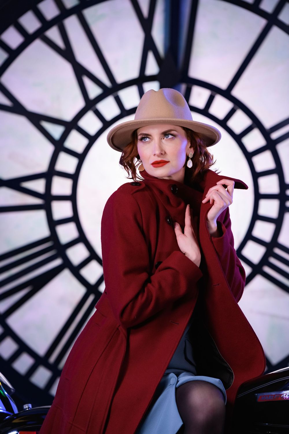 Elegant woman in red coat and fedora hat posing before large vintage clock.