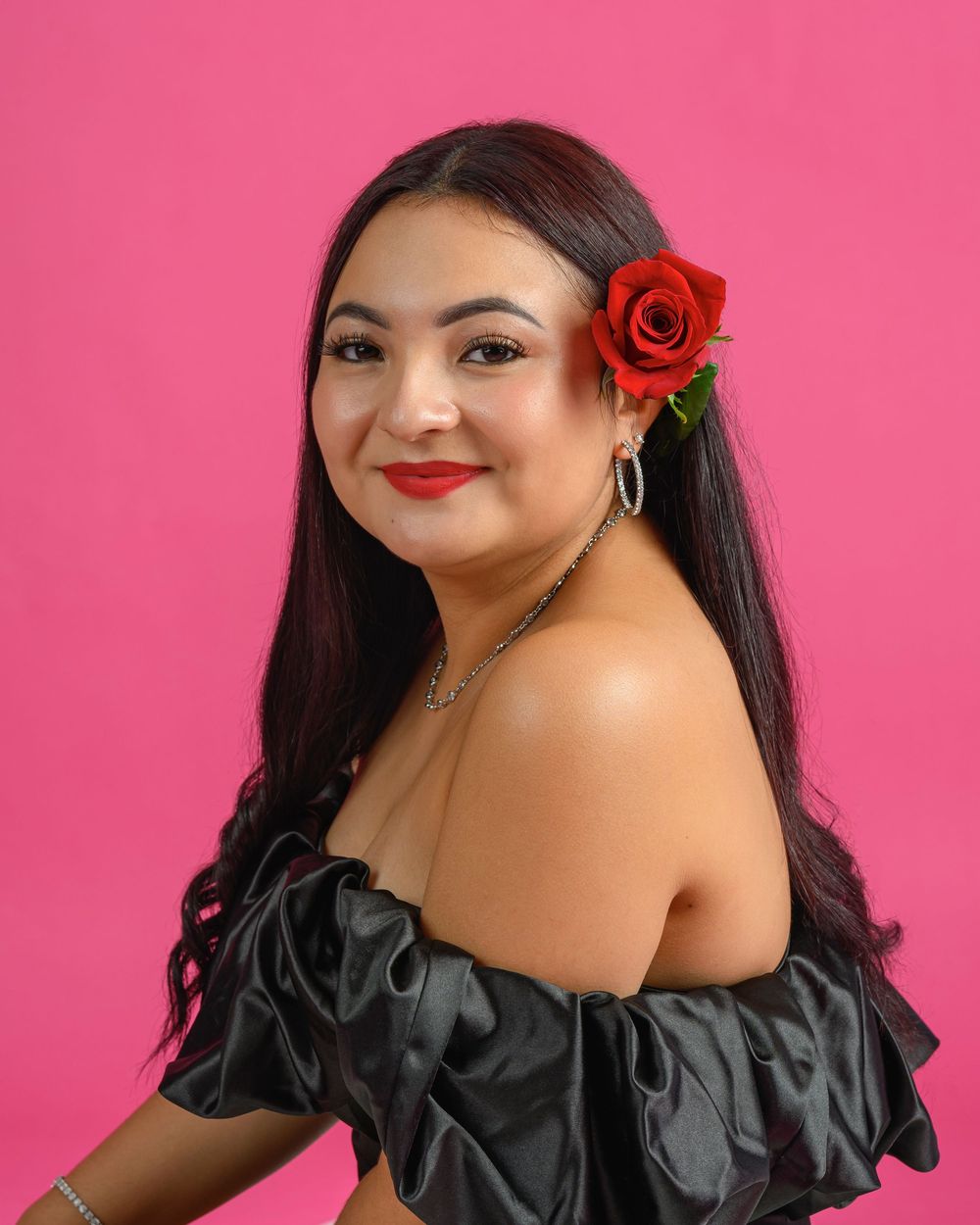 Young woman with red rose in dark hair wearing off-shoulder black dress on pink background.