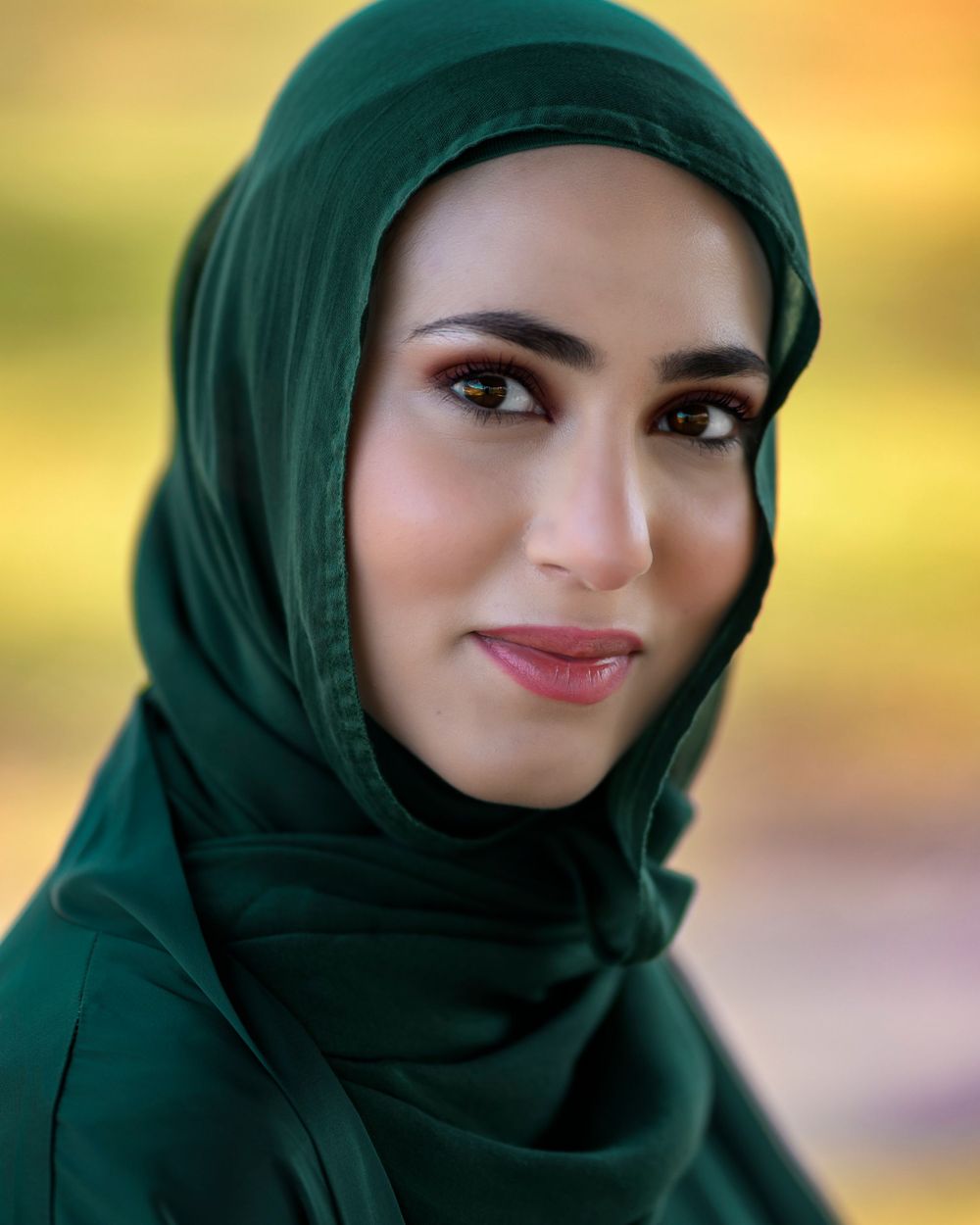 A beautiful woman wearing an elegant dark green hijab poses for a portrait outdoors.