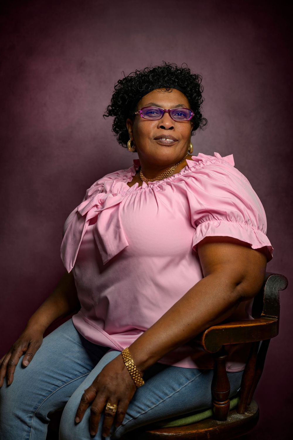 Woman in pink ruffled blouse seated in chair against purple studio background.