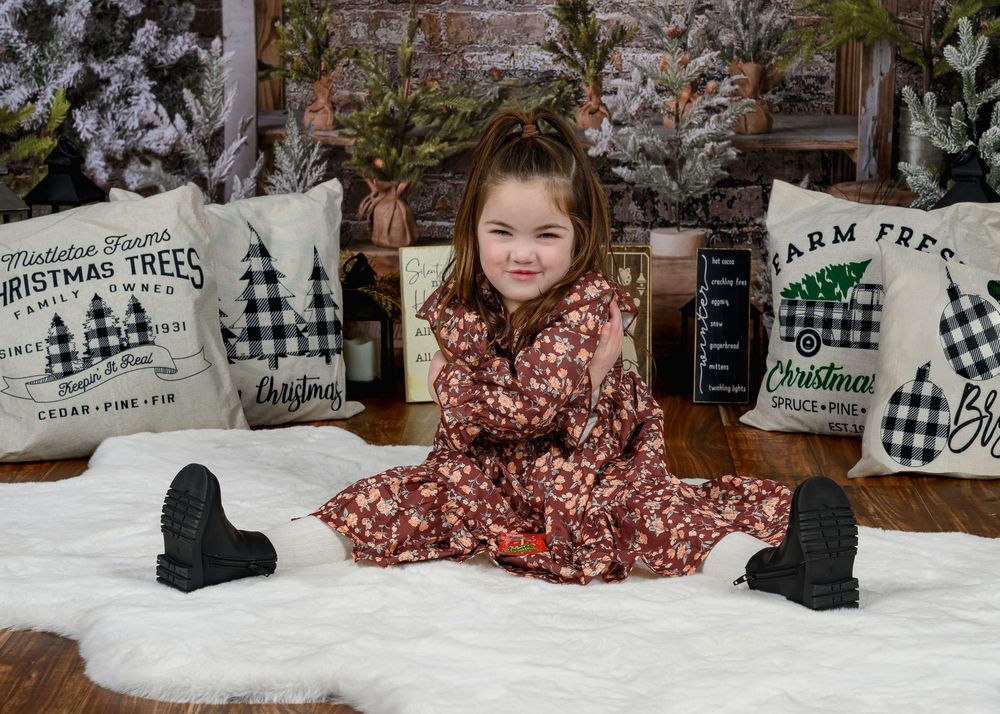 Young girl in floral dress sitting on white fur rug with Christmas decor.
