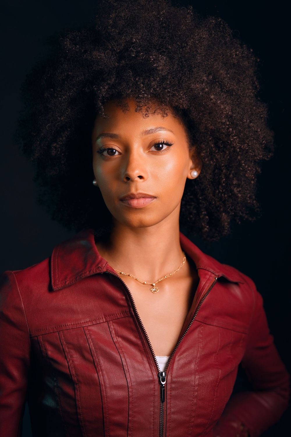 Young Black woman with natural afro hair wearing a red leather jacket and gold necklace against dark background.