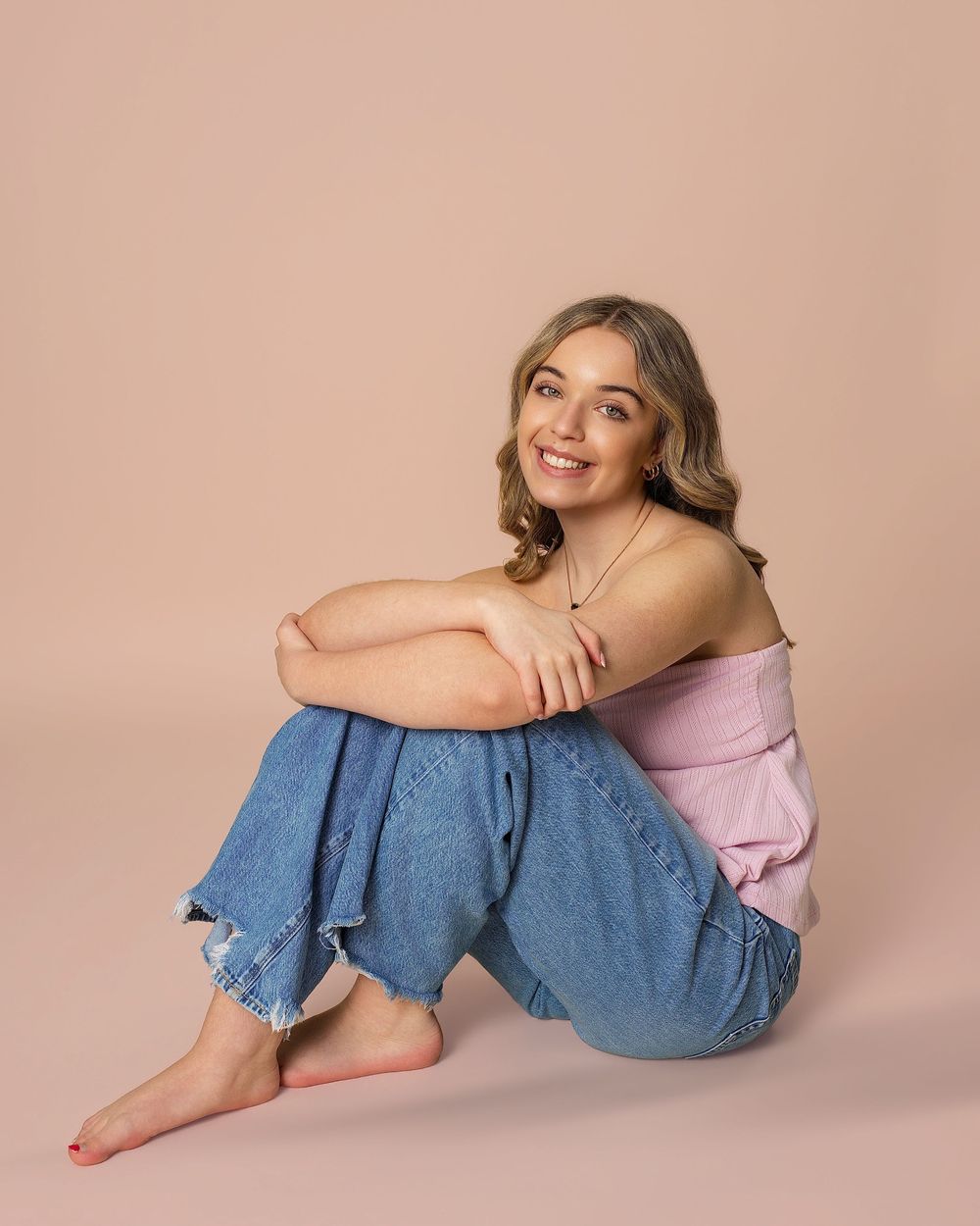 Smiling young blonde woman in pink top and wide-leg jeans sitting barefoot on beige studio floor.
Connecticut photographer