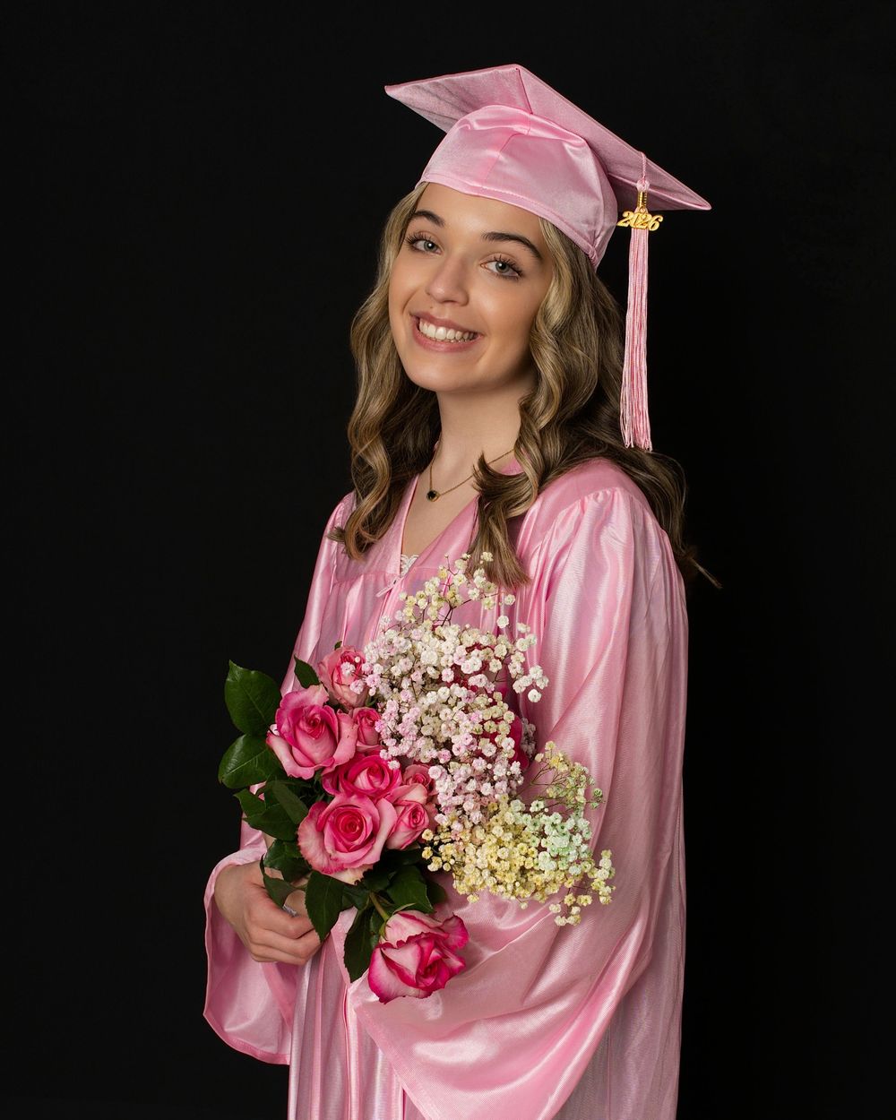 Smiling young woman in pink graduation cap and gown holding pink roses and baby's breath bouquet.