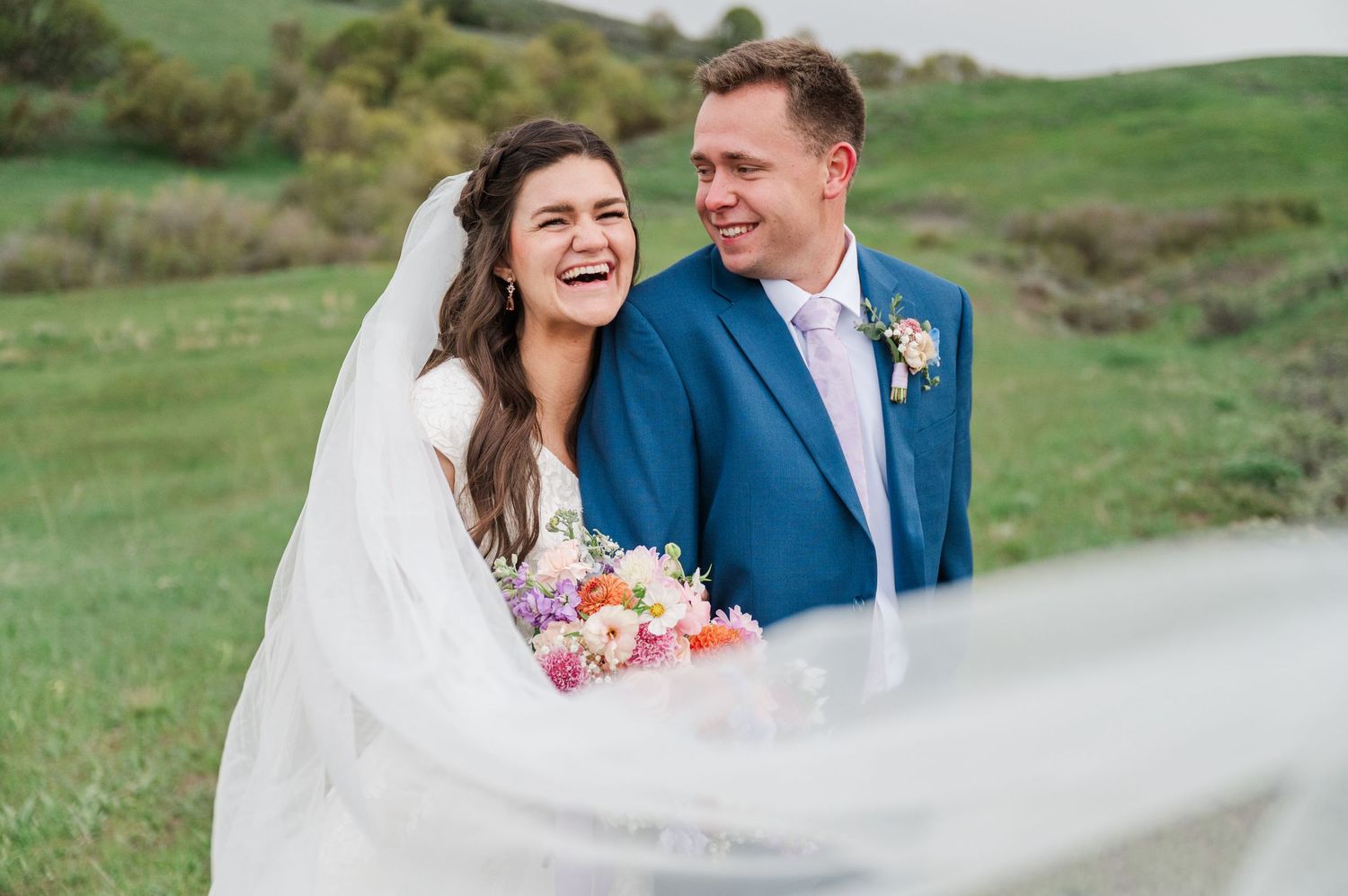Joyful wedding portrait with flowing veil against rolling green hills.