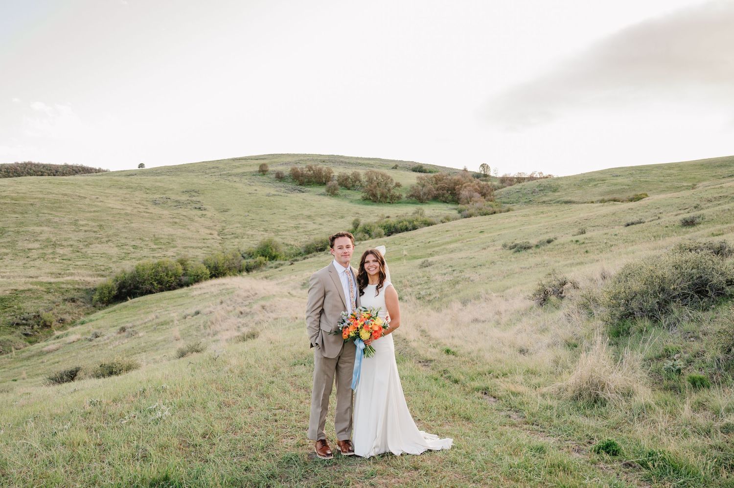 Bride and groom pose together on a lush green hillside, bride holding a vibrant bouquet on their wedding day.