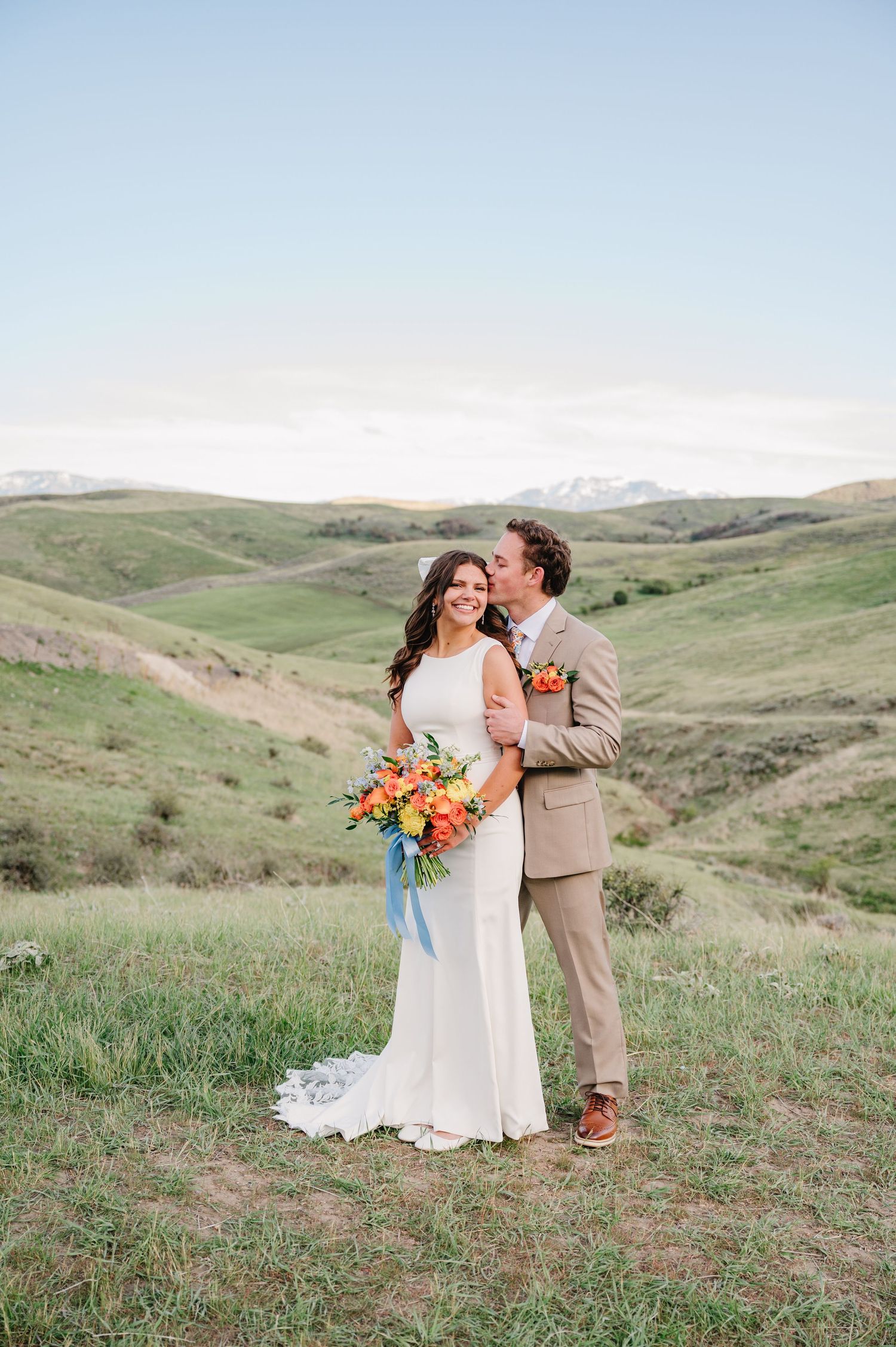 Bride and groom pose on green hillside, she holds vibrant orange bouquet with blue ribbons, snow-capped mountains in background.