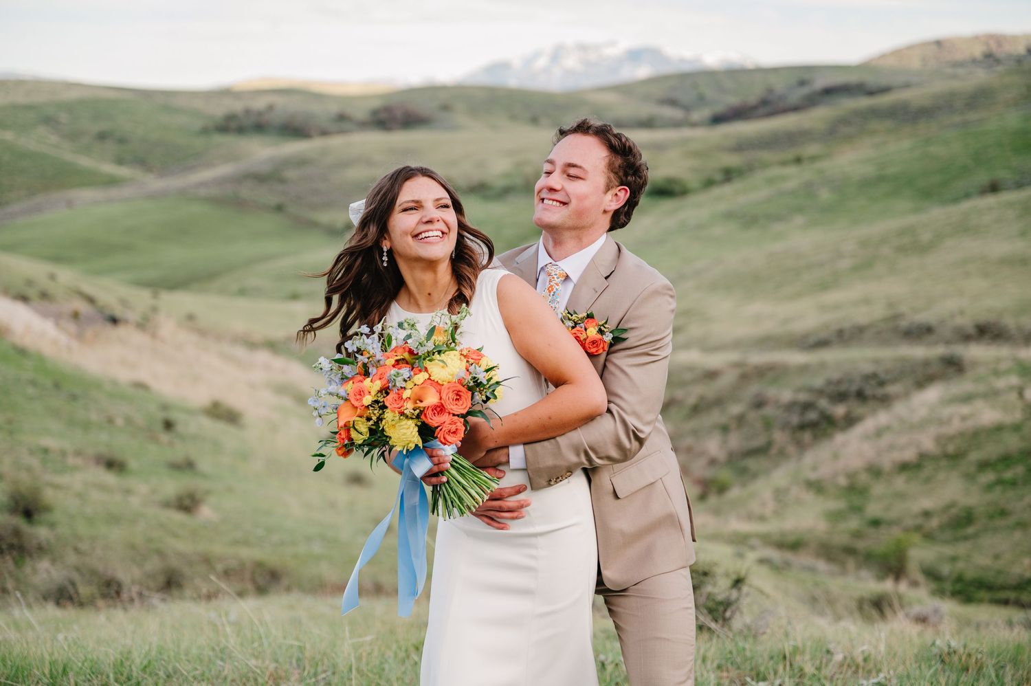 Bride and groom embrace in green rolling hills, she holds vibrant orange and yellow bouquet with blue ribbon.