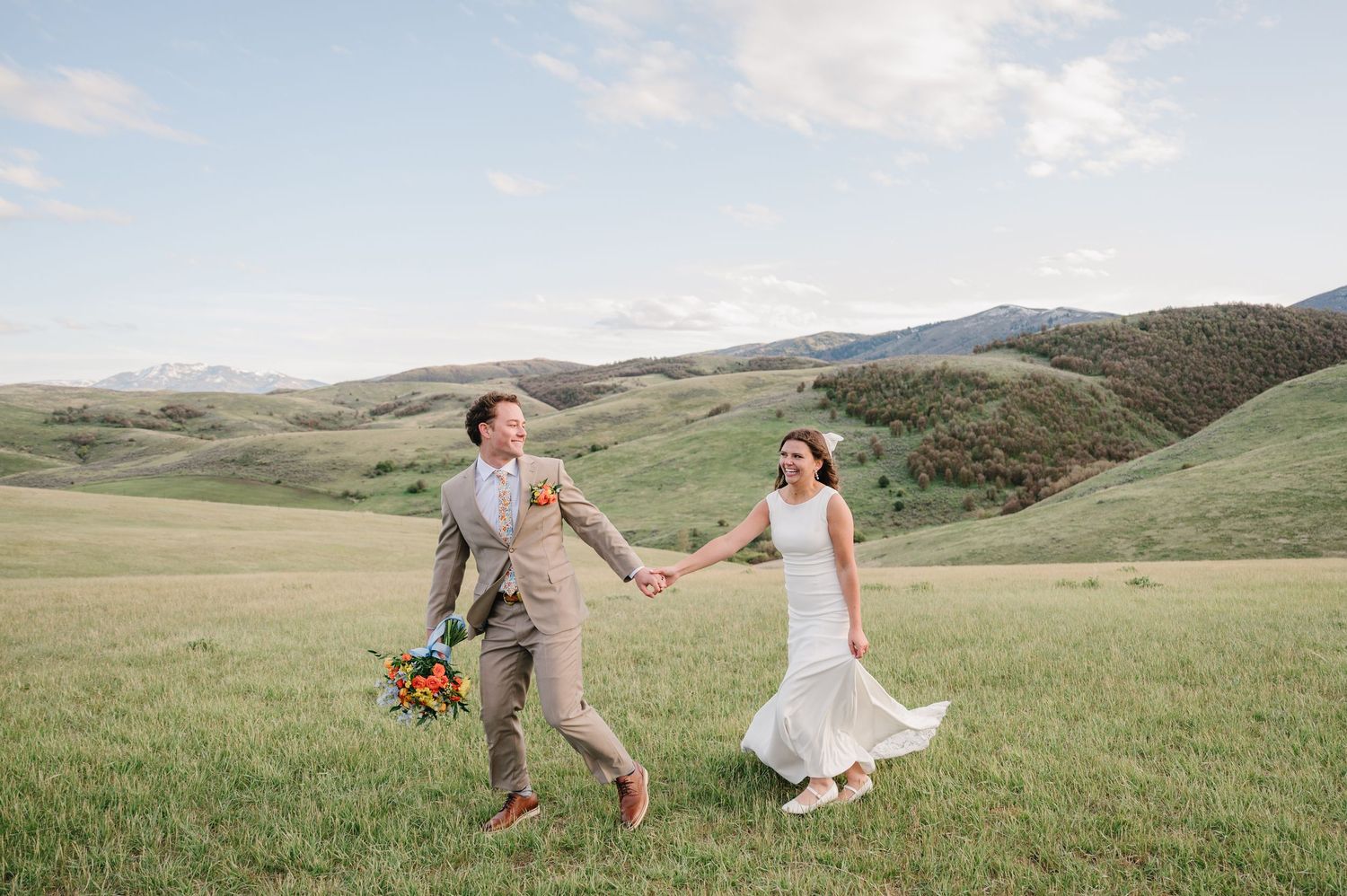 Bride and groom holding hands running through lush green meadow with rolling hills backdrop on wedding day.