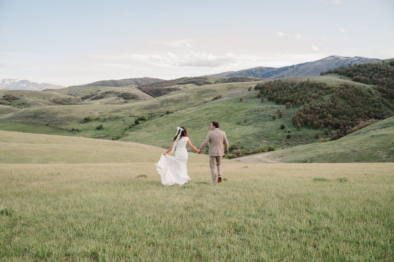 Bride and groom holding hands walking through lush green meadow with rolling hills and mountains in background.