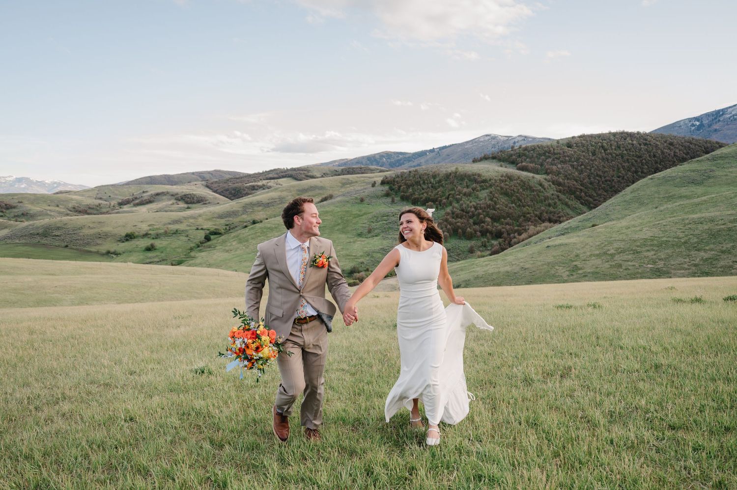 A joyful couple runs hand-in-hand through a lush green meadow with rolling hills, the bride in white and groom holding flowers.