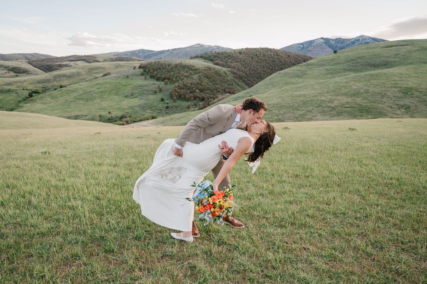 Bride and groom share a romantic dip kiss in a lush green meadow with rolling mountain hills in the background.