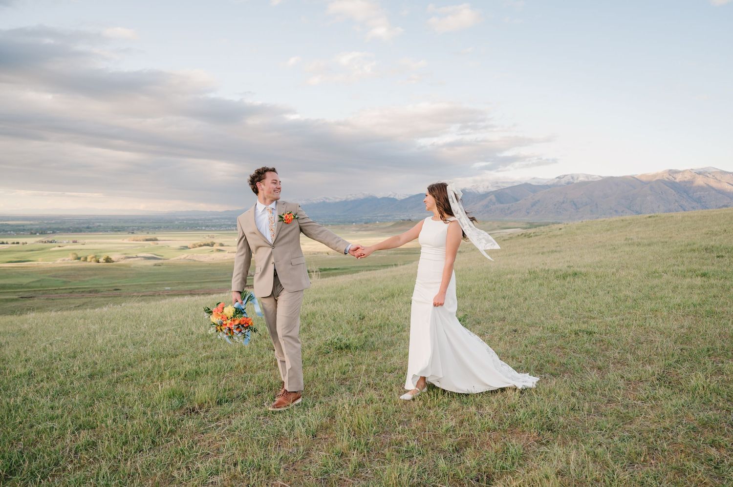 Bride and groom hold hands dancing in green mountain meadow, bride in white gown with veil, groom in tan suit holding bouquet.