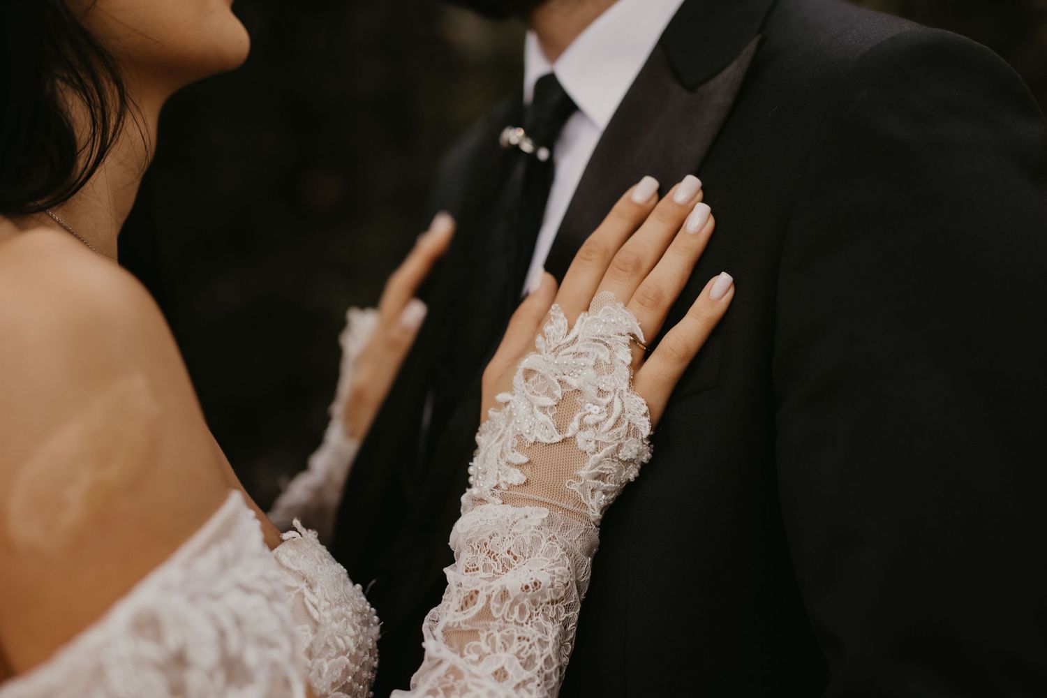 Close-up detail shot of a bride's lace sleeve and wedding ring against a dark suit.