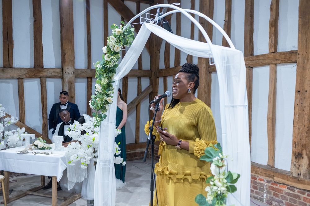 Speaker in yellow dress addresses wedding guests under a white floral arch.