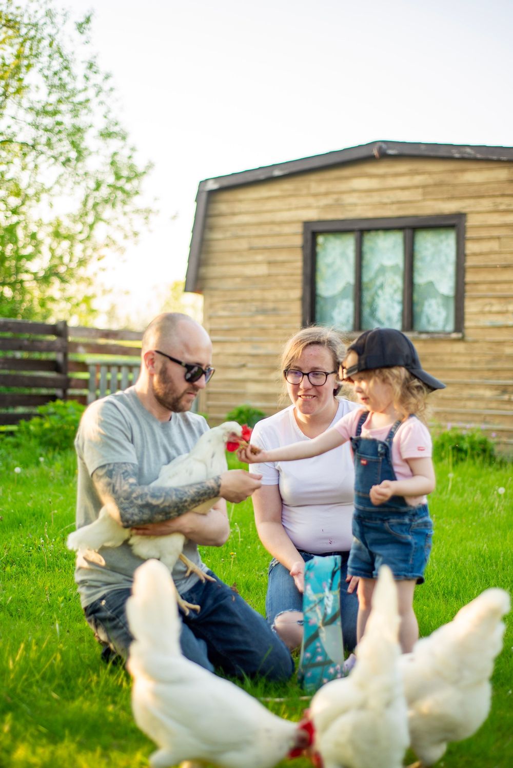 Family sits on grass near chickens outside their rustic home during a sunny afternoon.