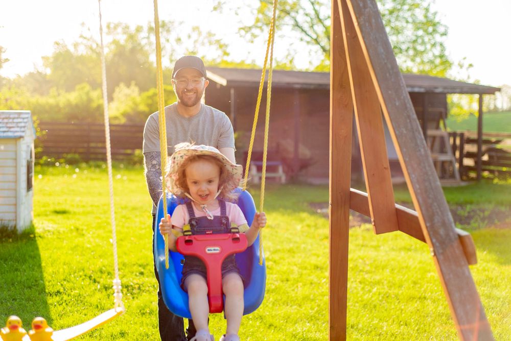 Child enjoying a pink toddler swing while someone pushes during golden hour in a backyard with green grass.