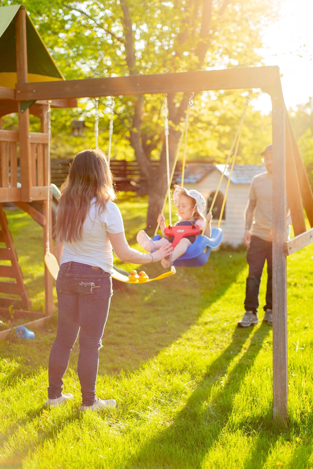 Family enjoying outdoor playtime at a wooden swing set during golden hour sunset in a backyard garden.