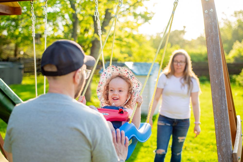 A happy child swings outside while a parent pushes and another adult stands nearby on a sunny afternoon.