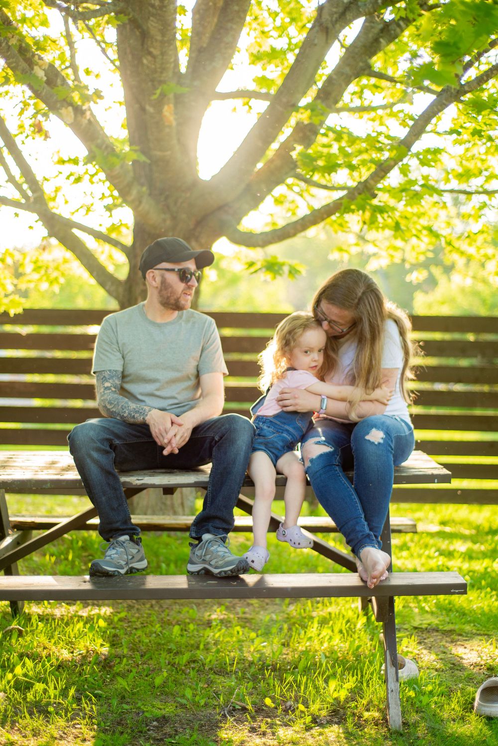 Family relaxing together on a wooden bench under a leafy tree in warm afternoon sunlight.