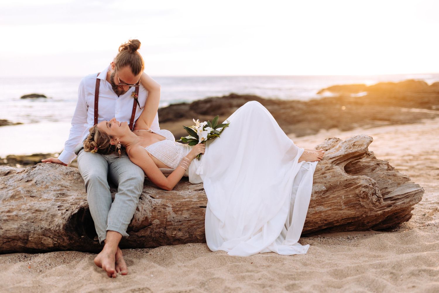 A couple rests on driftwood at the beach during their wedding photoshoot at sunset.
