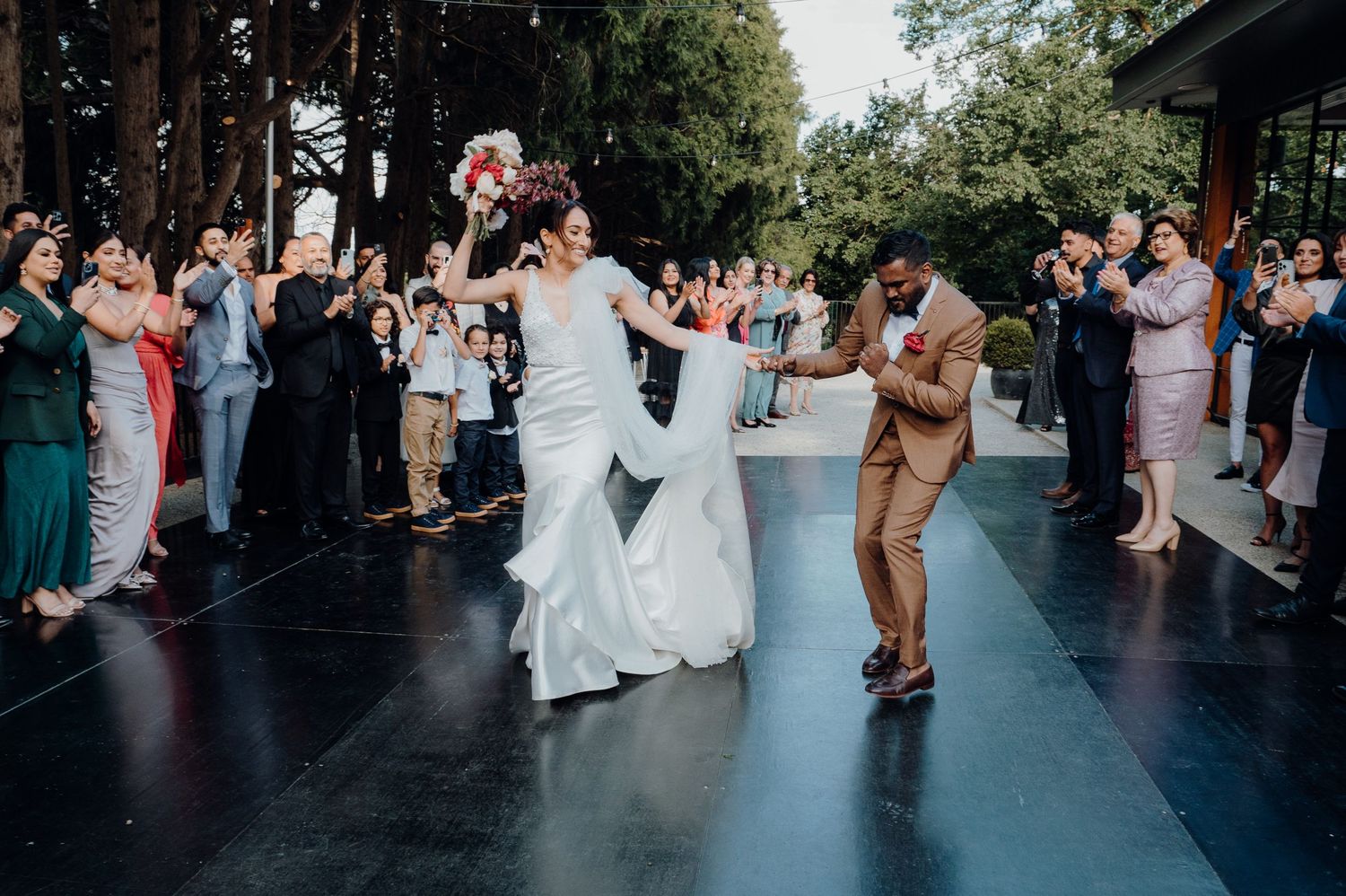 Wedding celebration with guests cheering as couple dances on outdoor black dancefloor.