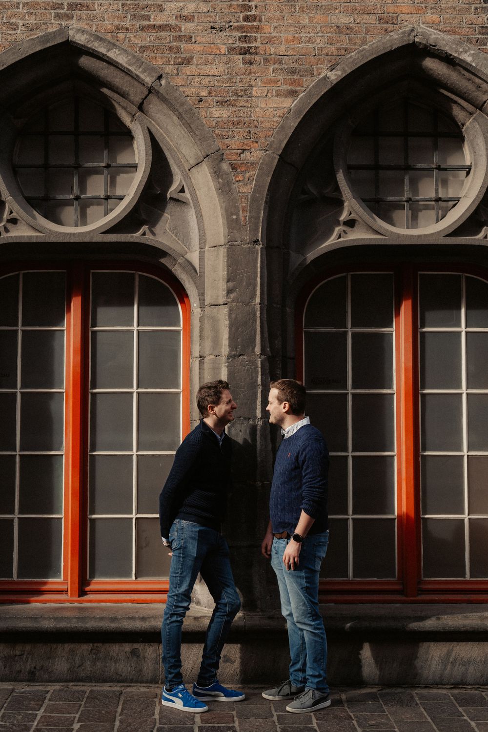 Two people in navy sweaters stand together against an architectural backdrop with arched windows and orange trim.