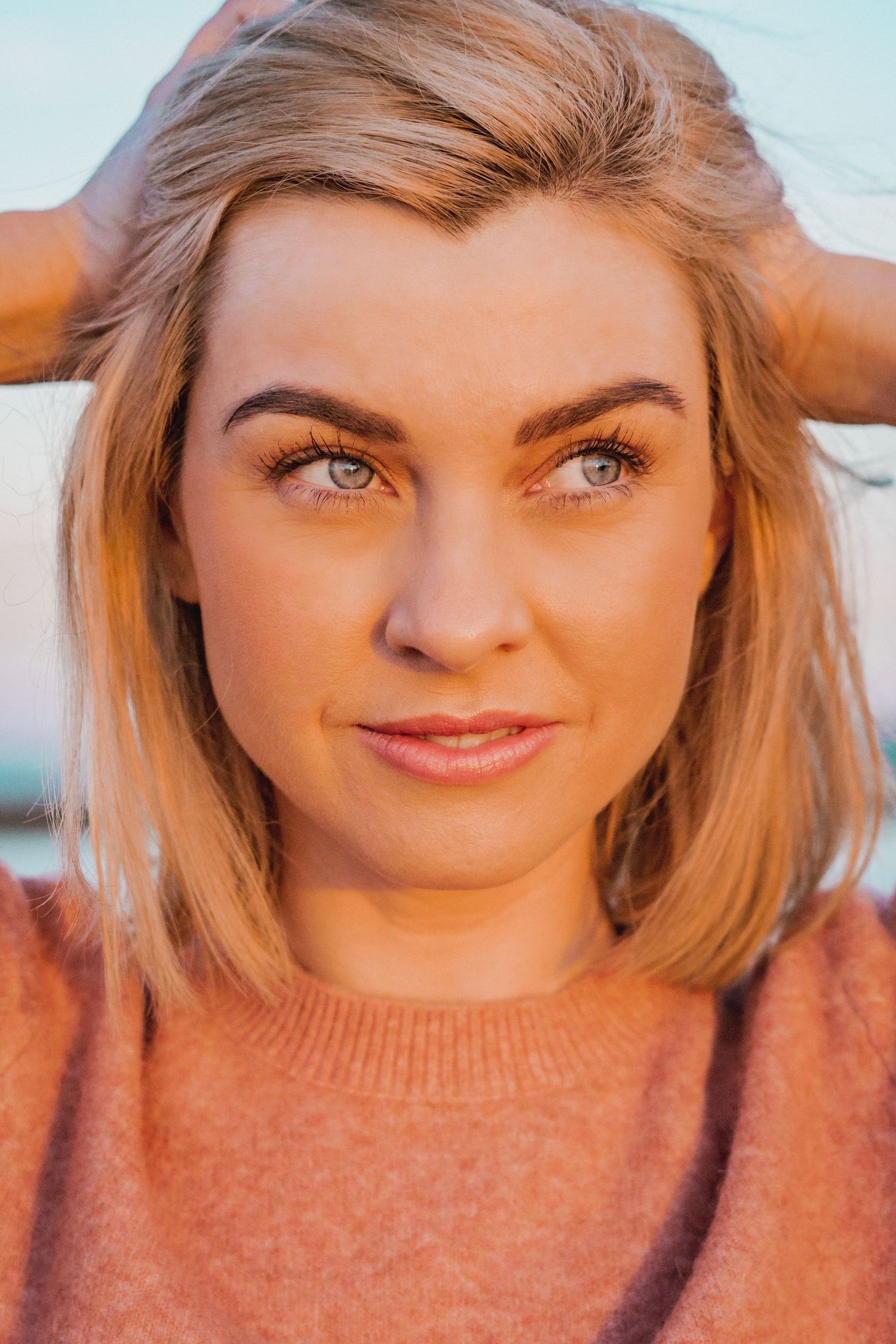 Close-up portrait of a blonde woman with blue eyes wearing a pink sweater outdoors.