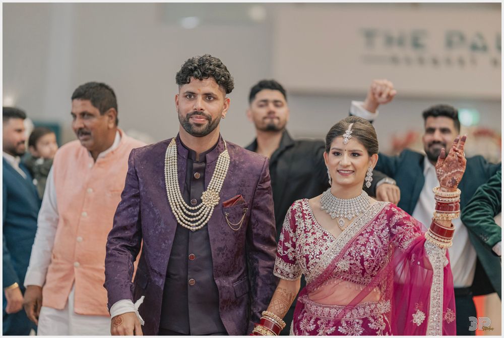 Couple in traditional Indian wedding attire walking with guests in background at indoor celebration.