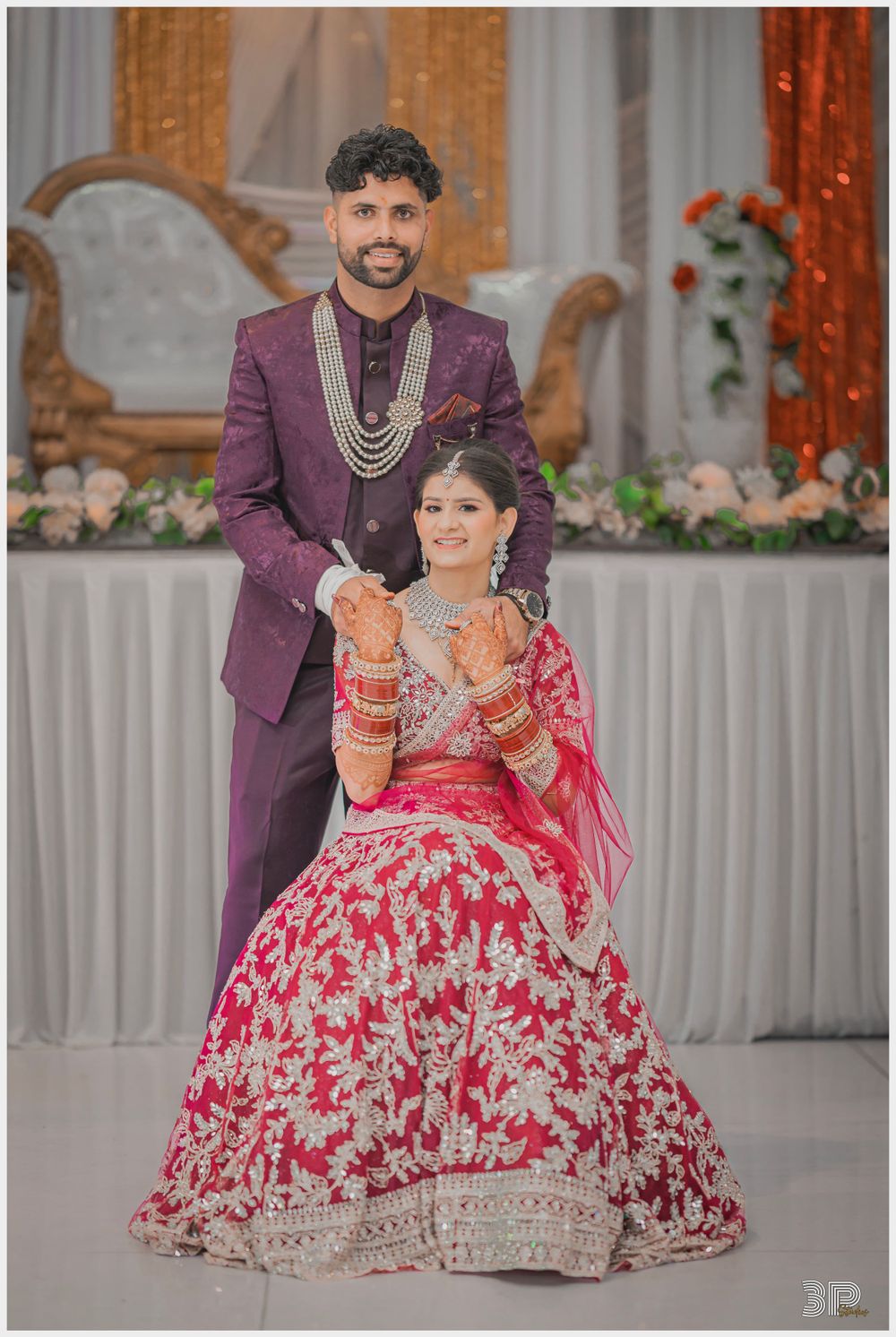 Wedding portrait of couple in traditional Indian attire against decorated backdrop with vintage sofa.