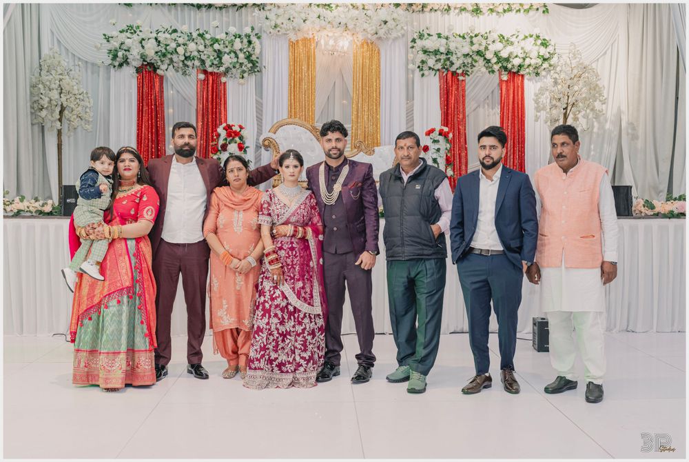 Wedding party group photo with colorful traditional outfits against decorated white and red backdrop.