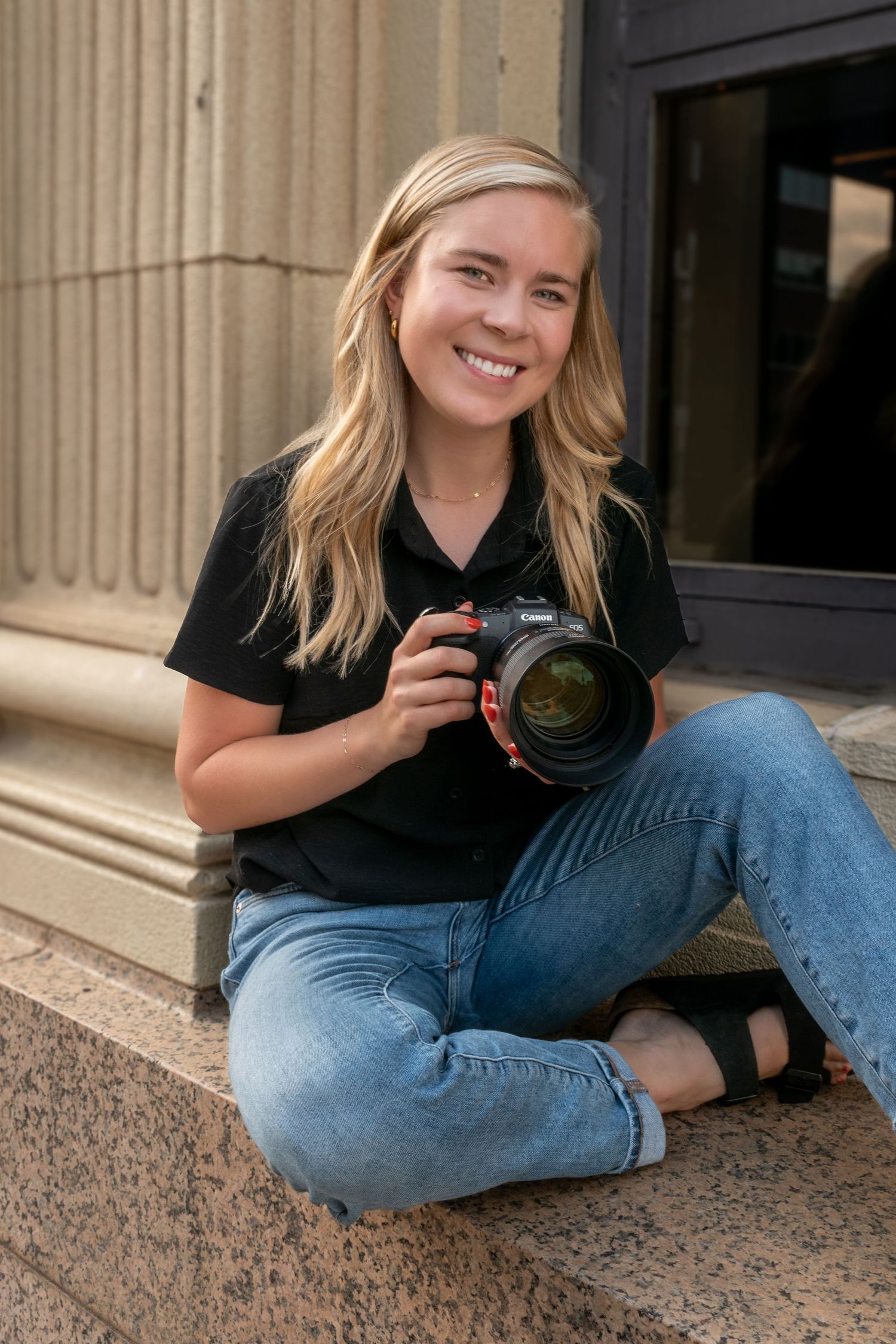 A blonde woman smiles while holding her own camera to the side of her face.