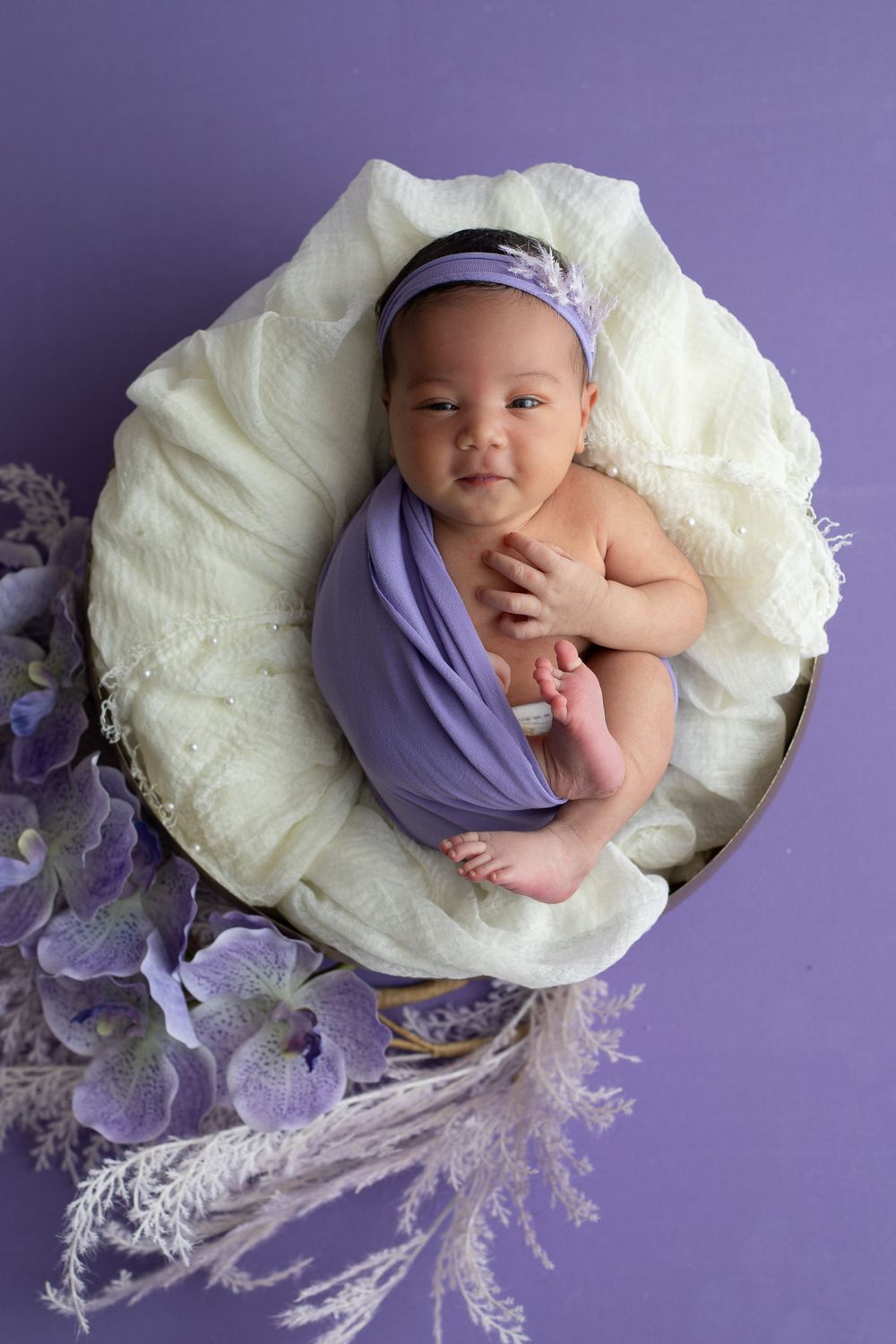 Newborn in purple wrap peacefully sleeping on white cushion with lavender floral decorations.